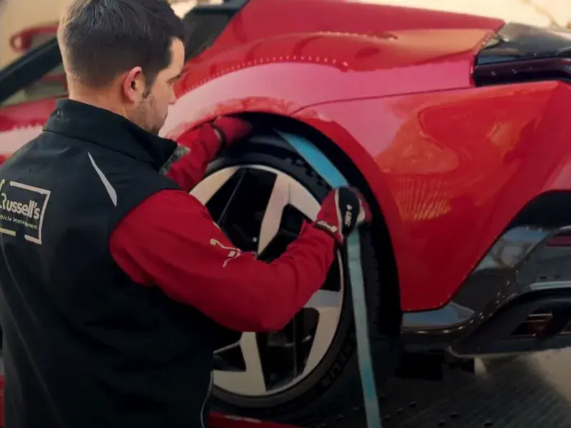 A worker in a black and red uniform secures a red sports car's wheel with a blue strap on a vehicle transport platform.