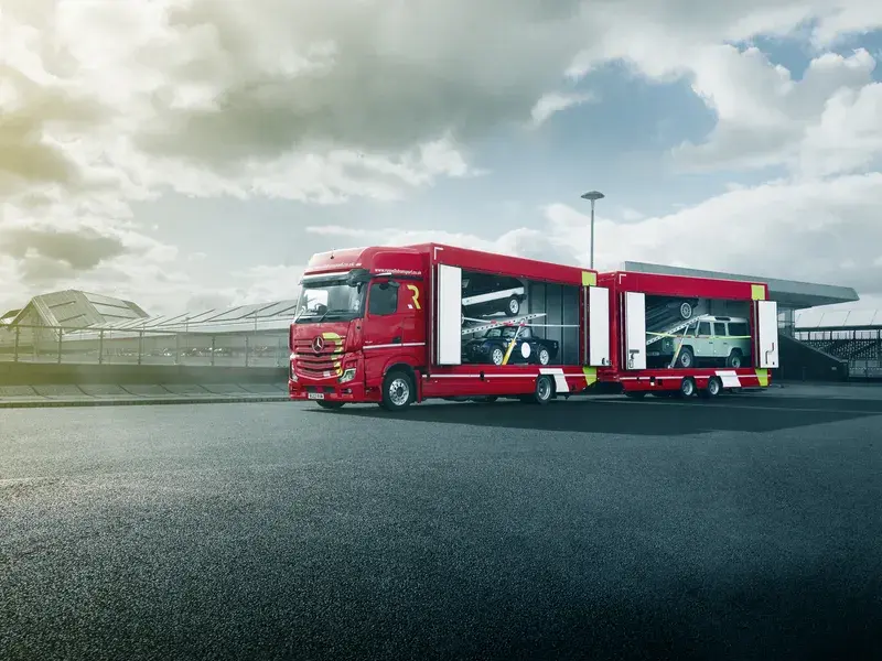 Red car transporter truck on a paved road under a cloudy sky, carrying two vintage cars, one green and one white.