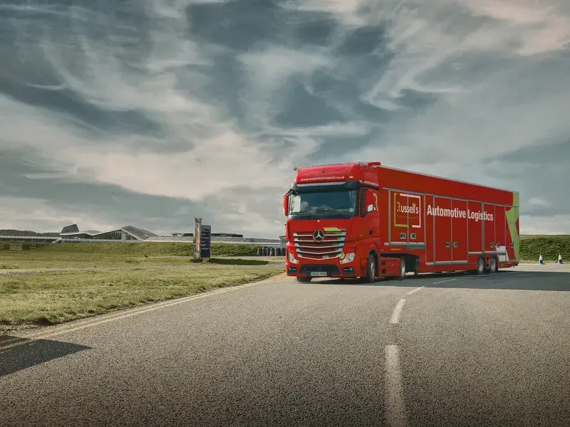 A red automotive logistics truck drives on a winding road with a grassy landscape and cloudy sky in the background.