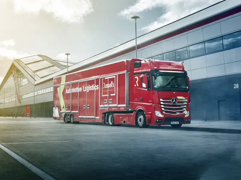 Red logistics truck parked beside a modern warehouse building under a blue sky, with sunlight streaming in from the left.