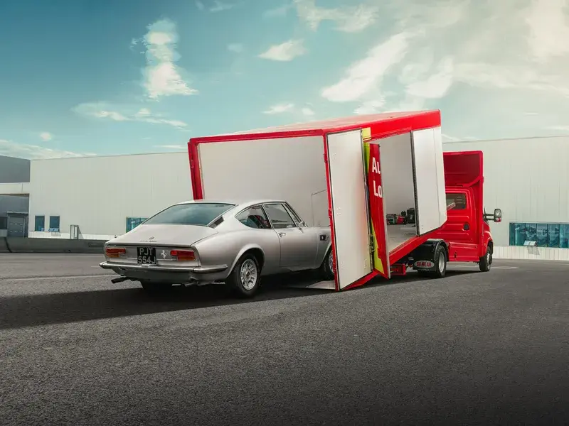 A classic silver car being loaded into a red enclosed car transporter on a sunny day, with industrial buildings in the background.