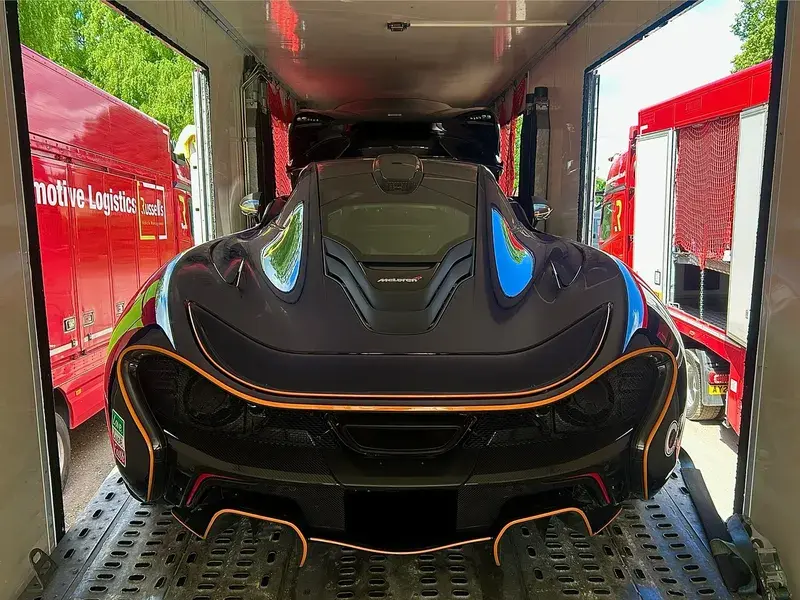 A sleek black Mclaren sports car with orange accents is secured inside a transport truck, surrounded by red vehicles and greenery outside.