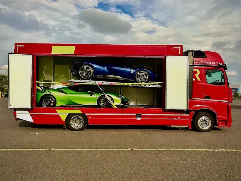 A red transport truck with open sides revealing two sports cars, one blue and one green, stacked inside.