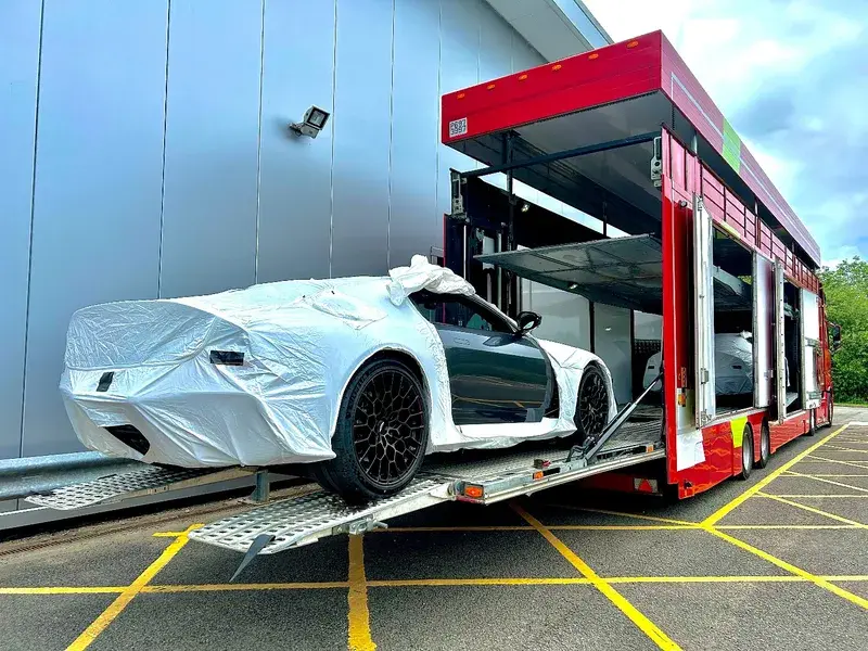 A sports car partially covered in protective wrap is being unloaded from a red transport truck near a large building.