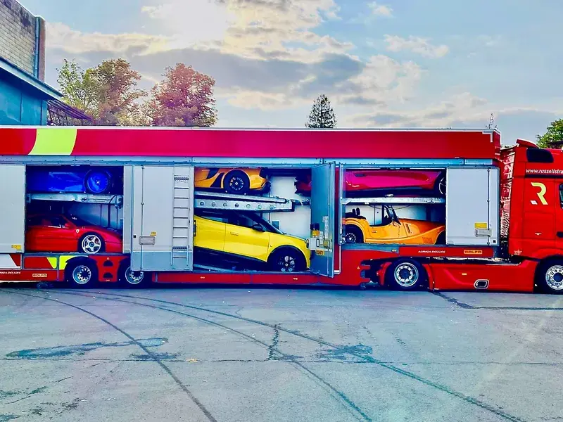 A red car carrier truck with open sides, revealing several brightly colored sports cars inside, parked on a paved area.